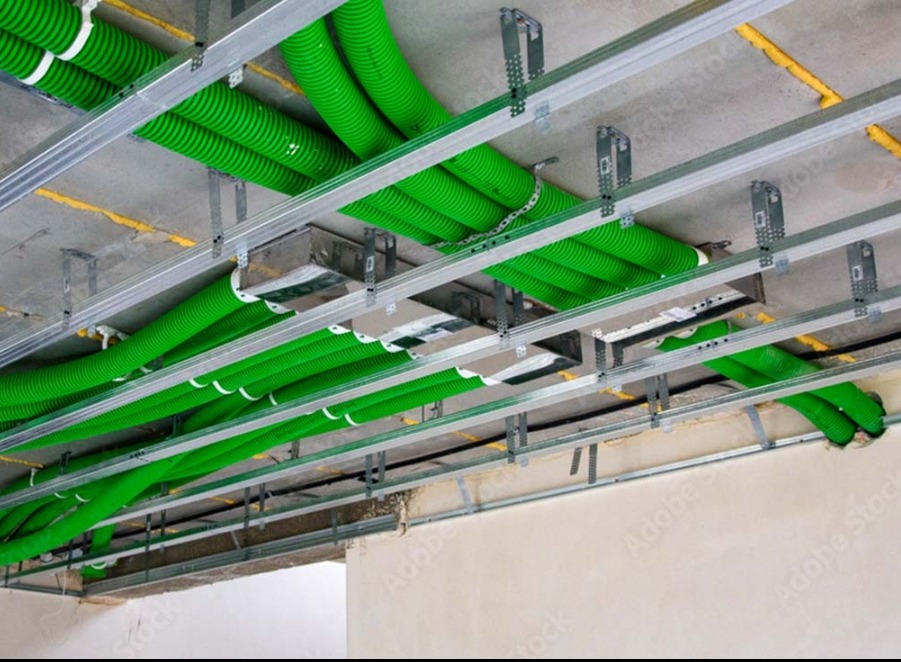Zielone, karbowane rury wentylacyjne zamocowane pod stropem za pomocą metalowych profili w surowym wnętrzu. Widoczne żółte uszczelnienia.