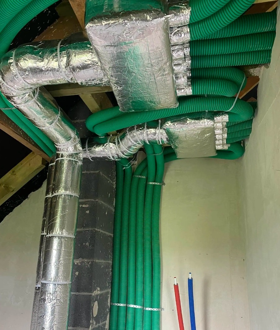 Zielone rury wentylacyjne i izolowane srebrną folią kanały w budynku, zamocowane do sufitu i ściany. Elementy instalacji rekuperacji.