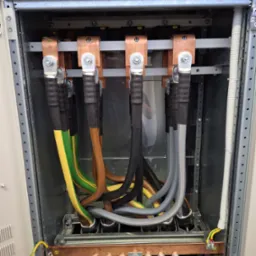 Rozdzielnica elektryczna z miedzianymi szynami, oznaczonymi L1, L2, L3, oraz grubymi kablami w izolacji, w kolorach: żółto-zielonym, brązowym, czarnym i szarym.