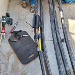 ElektroInstalMontaz - Przygotowane kable elektryczne w czarnej izolacji, narzędzia pracy elektryka oraz podkładka marki Wurth na betonowym podłożu. Widoczne buty i fragmenty odzieży roboczej.