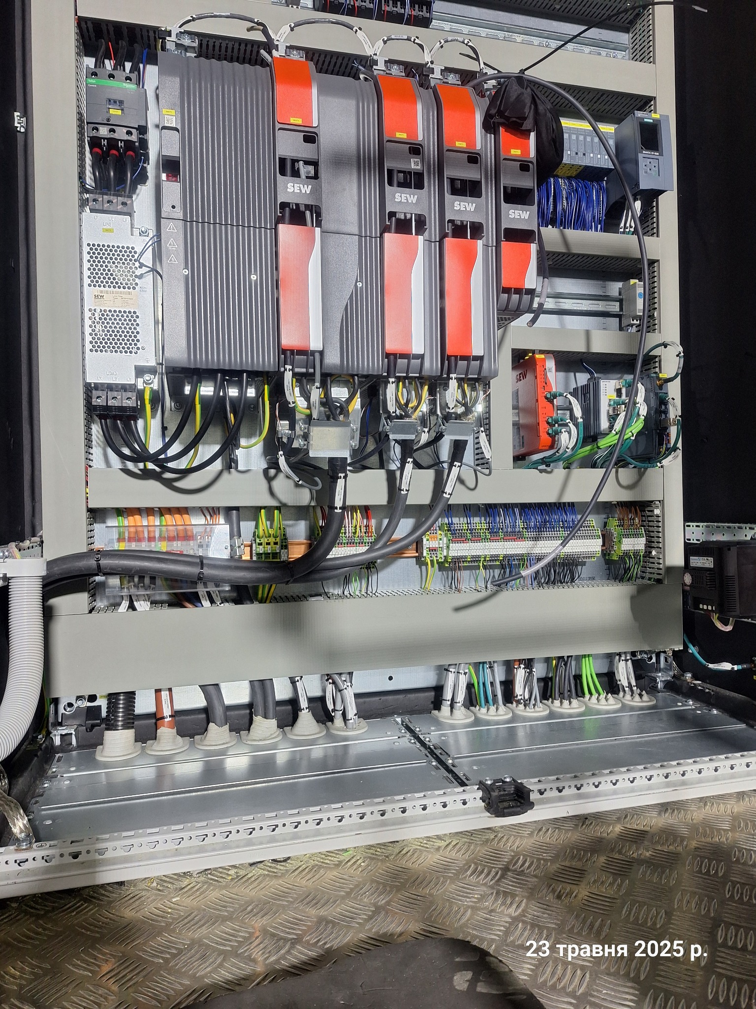 Rozbudowana szafa sterownicza z modułami SEW, licznymi przewodami i elementami elektrycznymi, widok z dołu na metalową konstrukcję i podłogę z blachy ryflowanej.