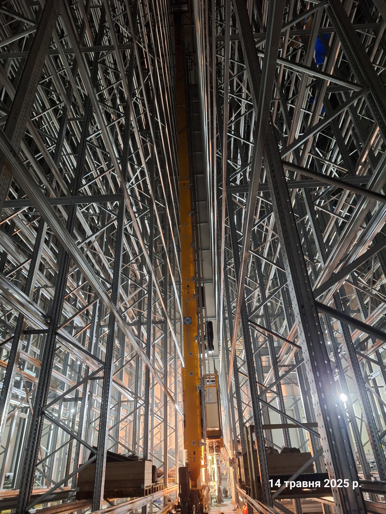 Wysokie, metalowe regały magazynowe z żółtym wózkiem widłowym w centrum, oświetlone od dołu. Perspektywa z dołu do góry podkreśla skalę konstrukcji.