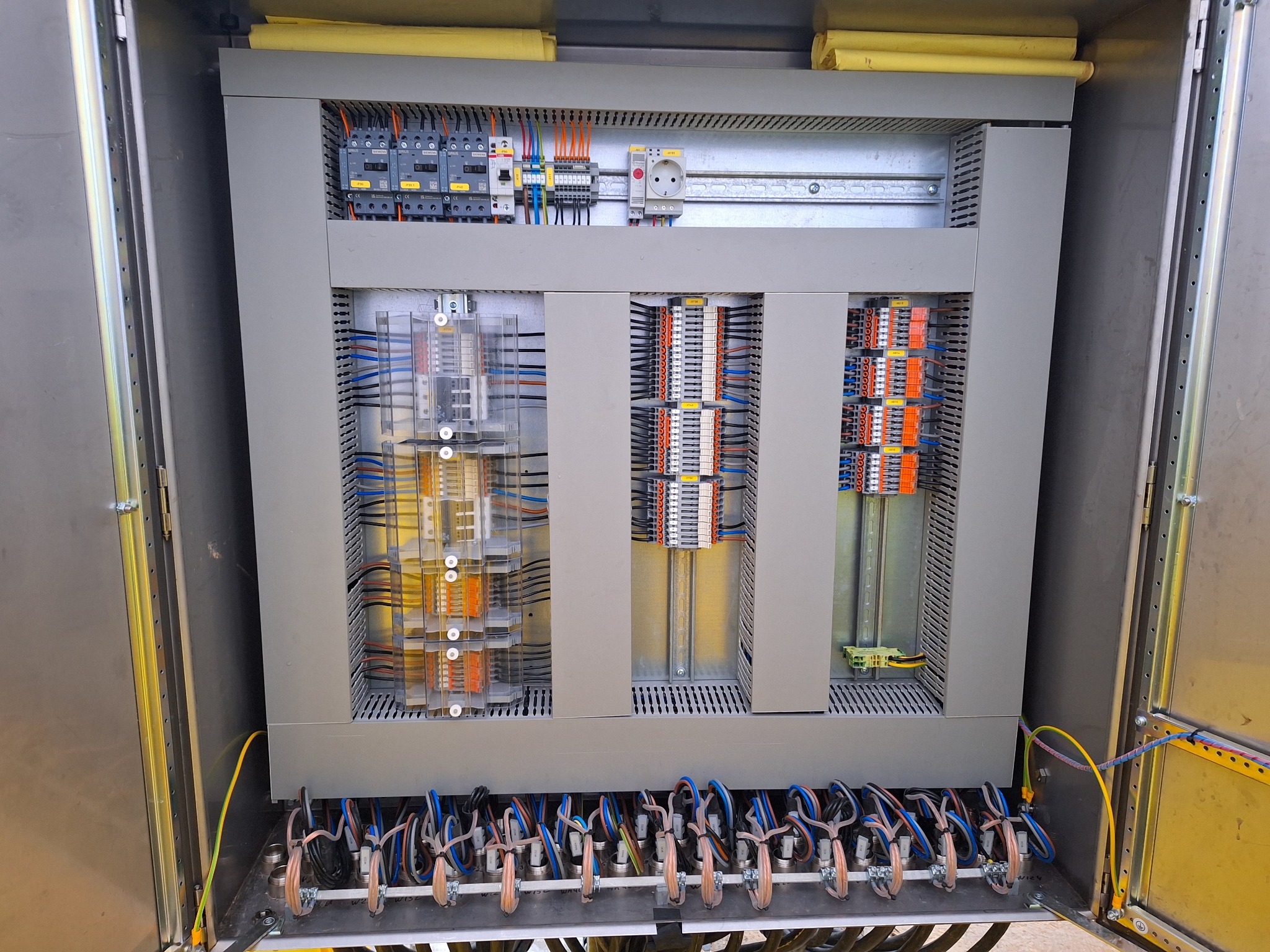 Otwarta szafa sterownicza z precyzyjnie ułożonymi kablami, widoczne moduły sterujące i złącza, całość w szarej obudowie z metalowymi elementami.