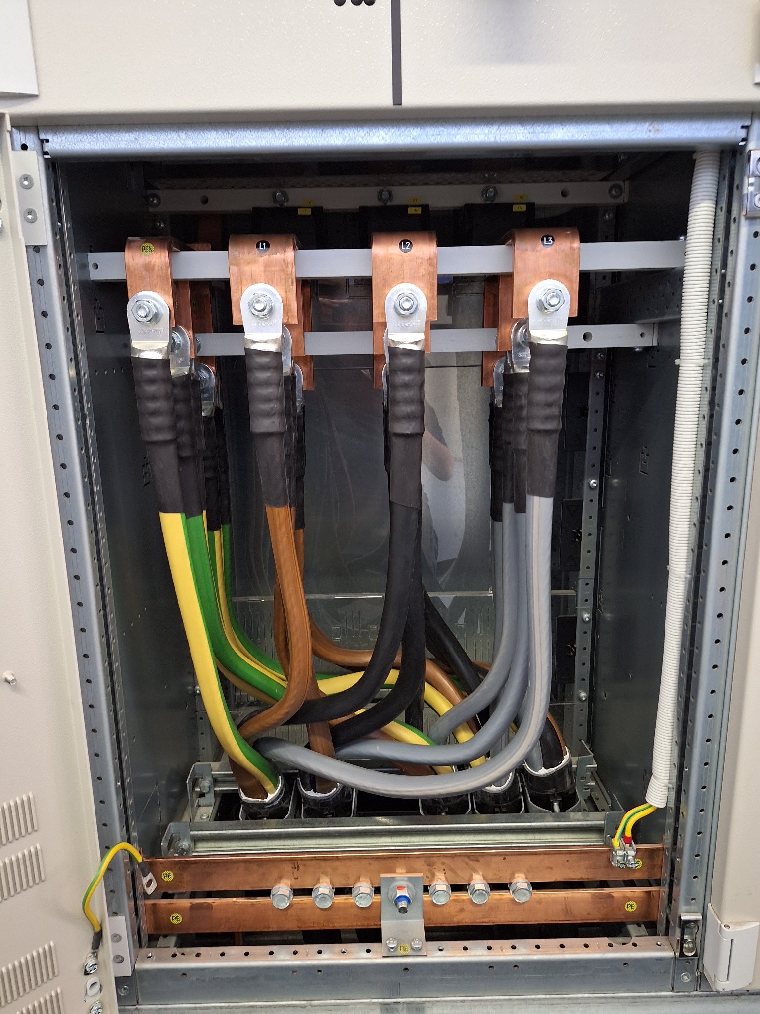 Rozdzielnica elektryczna z miedzianymi szynami, oznaczonymi L1, L2, L3, oraz grubymi kablami w izolacji, w kolorach: żółto-zielonym, brązowym, czarnym i szarym.