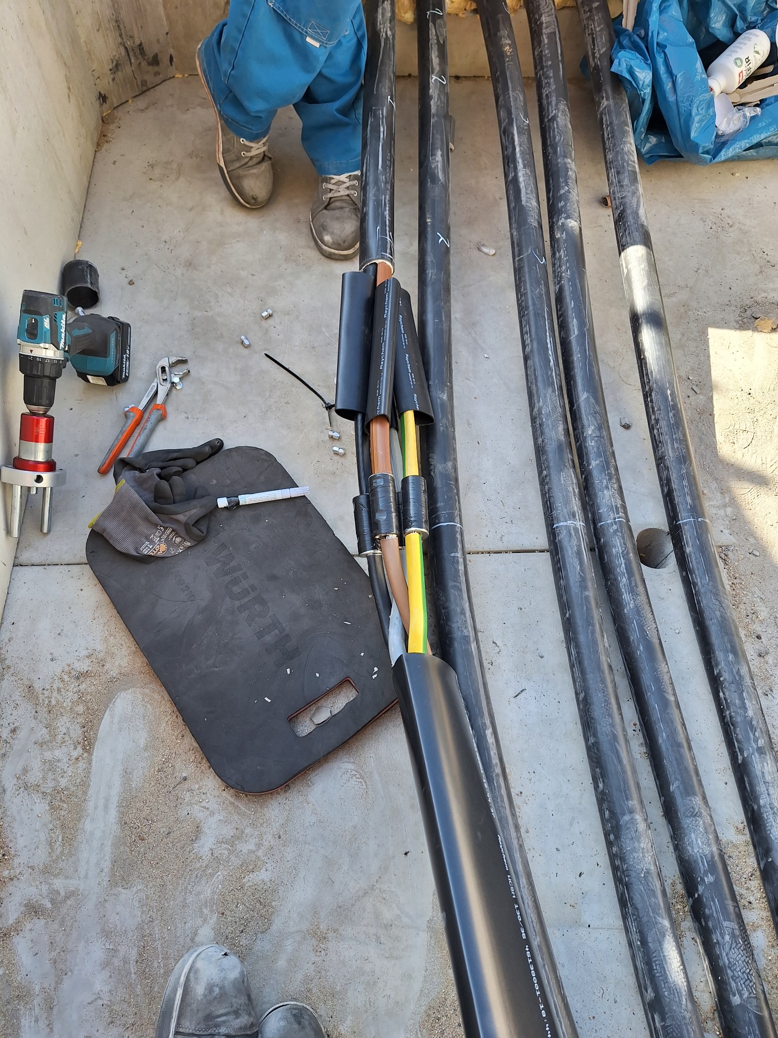 Przygotowane kable elektryczne w czarnej izolacji, narzędzia pracy elektryka oraz podkładka marki Wurth na betonowym podłożu. Widoczne buty i fragmenty odzieży roboczej.