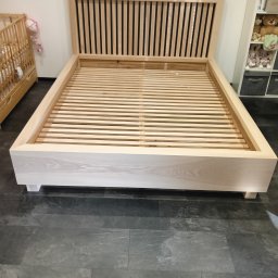 Schody Damian Szpak Zakład Stolarski - Jasne, drewniane łóżko z listwowym zagłówkiem, ustawione na ciemnej podłodze. Widoczny fragment dziecięcego łóżeczka i regał z zabawkami w tle.