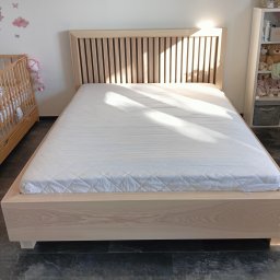 Schody Damian Szpak Zakład Stolarski - Drewniane łóżko z zagłówkiem z pionowych listew w jasnym odcieniu drewna. Obok łóżeczko dziecięce i regał z zabawkami. Delikatne oświetlenie podkreśla naturalne wykończenie.