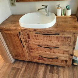 Schody Damian Szpak Zakład Stolarski - Drewniana szafka łazienkowa z umywalką nablatową. Front z trzema szufladami, dekoracyjne uchwyty. Blat z widoczną strukturą drewna. Styl rustykalny.