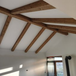 Schody Damian Szpak Zakład Stolarski - Jasne wnętrze poddasza z widocznymi, dekoracyjnymi belkami stropowymi z ciemnego drewna. W tle okno i fragment instalacji kominowej.