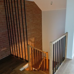 Schody Damian Szpak Zakład Stolarski - Nowoczesne schody drewniane z czarną, metalową balustradą na tle ceglanej ściany. Jasne drewno schodów kontrastuje z ciemnymi elementami balustrady.