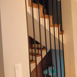 Nowoczesna balustrada metalowa z pionowymi prętami, przez którą widać fragment drewnianych schodów. Minimalistyczny design, jasne wnętrze, widoczny włącznik światła.