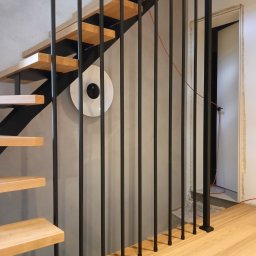 Schody Damian Szpak Zakład Stolarski - Nowoczesne schody z drewnianymi stopniami i czarną, metalową balustradą z pionowych prętów. Na ścianie okrągła lampa. Widok na fragment podłogi i otwarte drzwi.