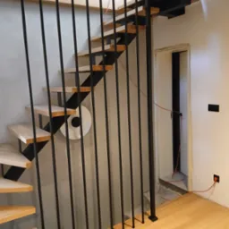 Nowoczesne schody z drewnianymi stopniami i czarną, metalową balustradą w minimalistycznym wnętrzu. Widoczne detale wykończenia i oświetlenie.