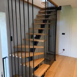 Nowoczesne schody z drewnianymi stopniami i czarną, metalową balustradą z pionowych prętów. Jasne drewno, minimalistyczny design, widok z dołu.