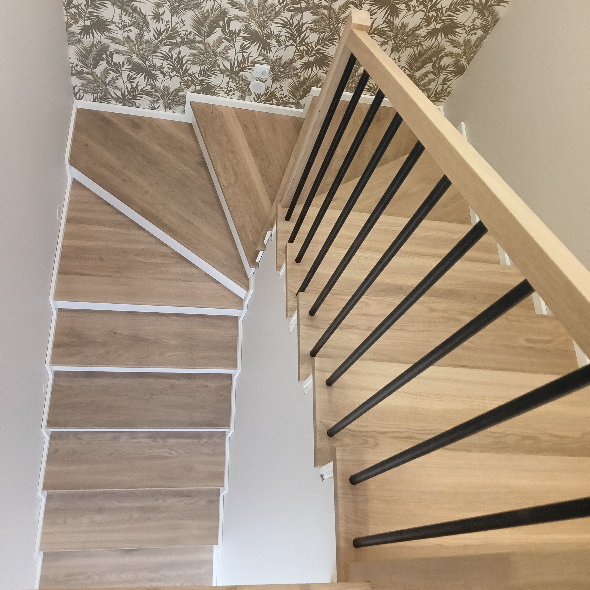 Nowoczesne schody z jasnego drewna z czarną balustradą, widok z góry, na tle ściany z tapetą w liście. Minimalistyczny design i precyzyjne wykonanie.