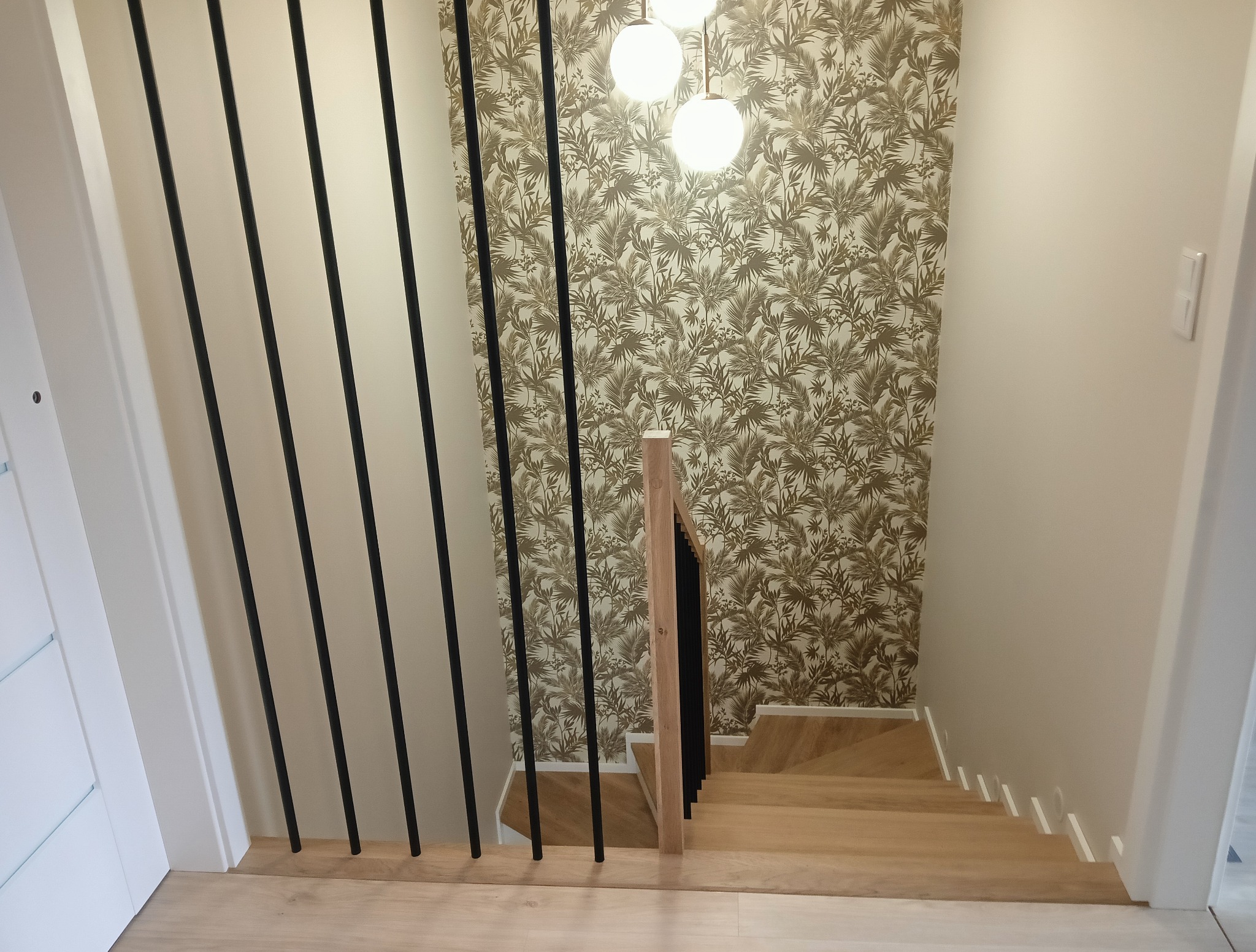 Jasne schody z drewnianymi stopniami i czarnymi, pionowymi balustradami na tle ściany w tropikalne wzory, oświetlone trzema kulistymi lampami.
