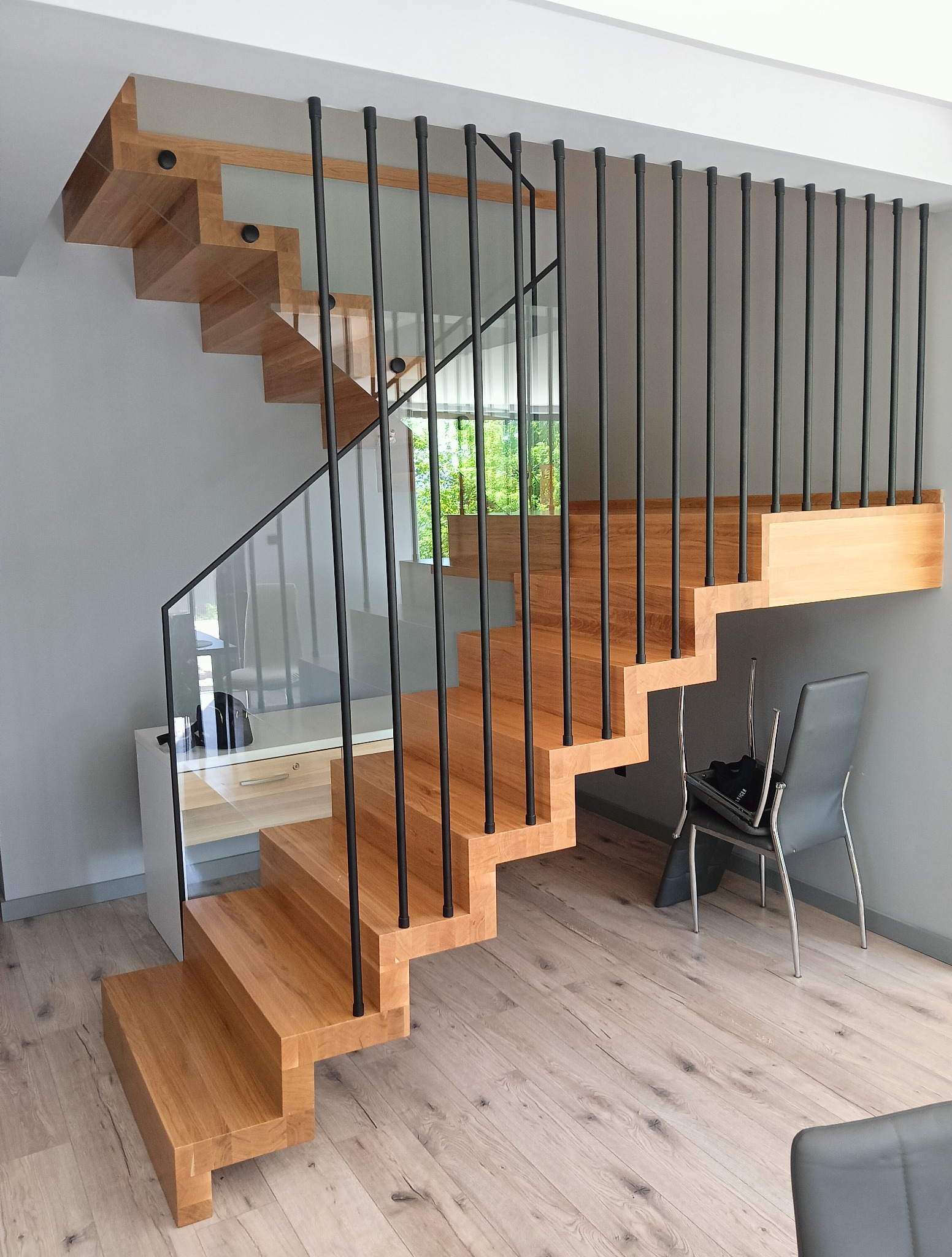 Nowoczesne schody drewniane z czarnymi, metalowymi balustradami i szklanym panelem bocznym. Naturalne oświetlenie, widok na zieleń za oknem.