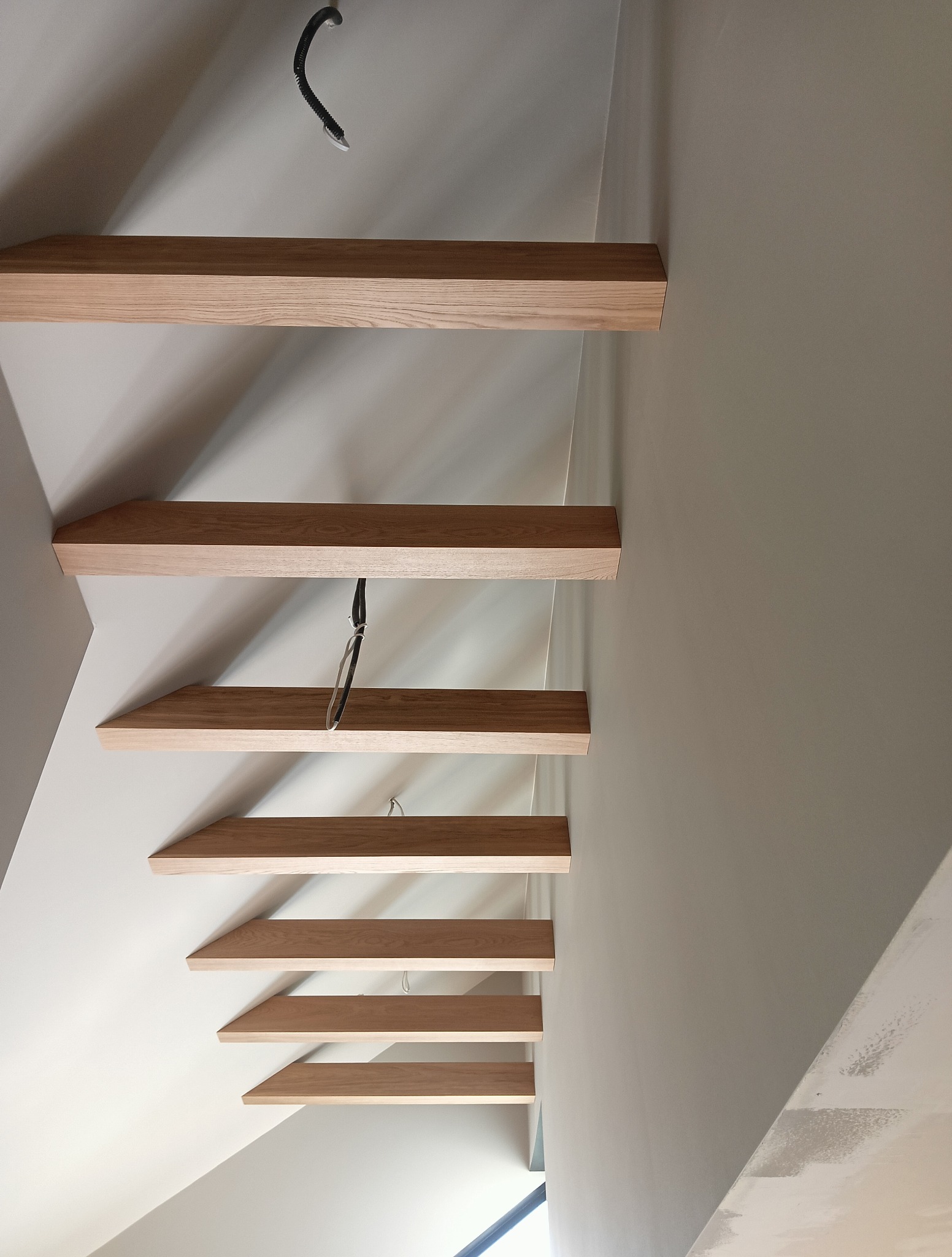 Minimalistyczne, wiszące schody drewniane o jasnym odcieniu, zamocowane do ściany. Widok z dołu, ukazujący konstrukcję i oświetlenie.