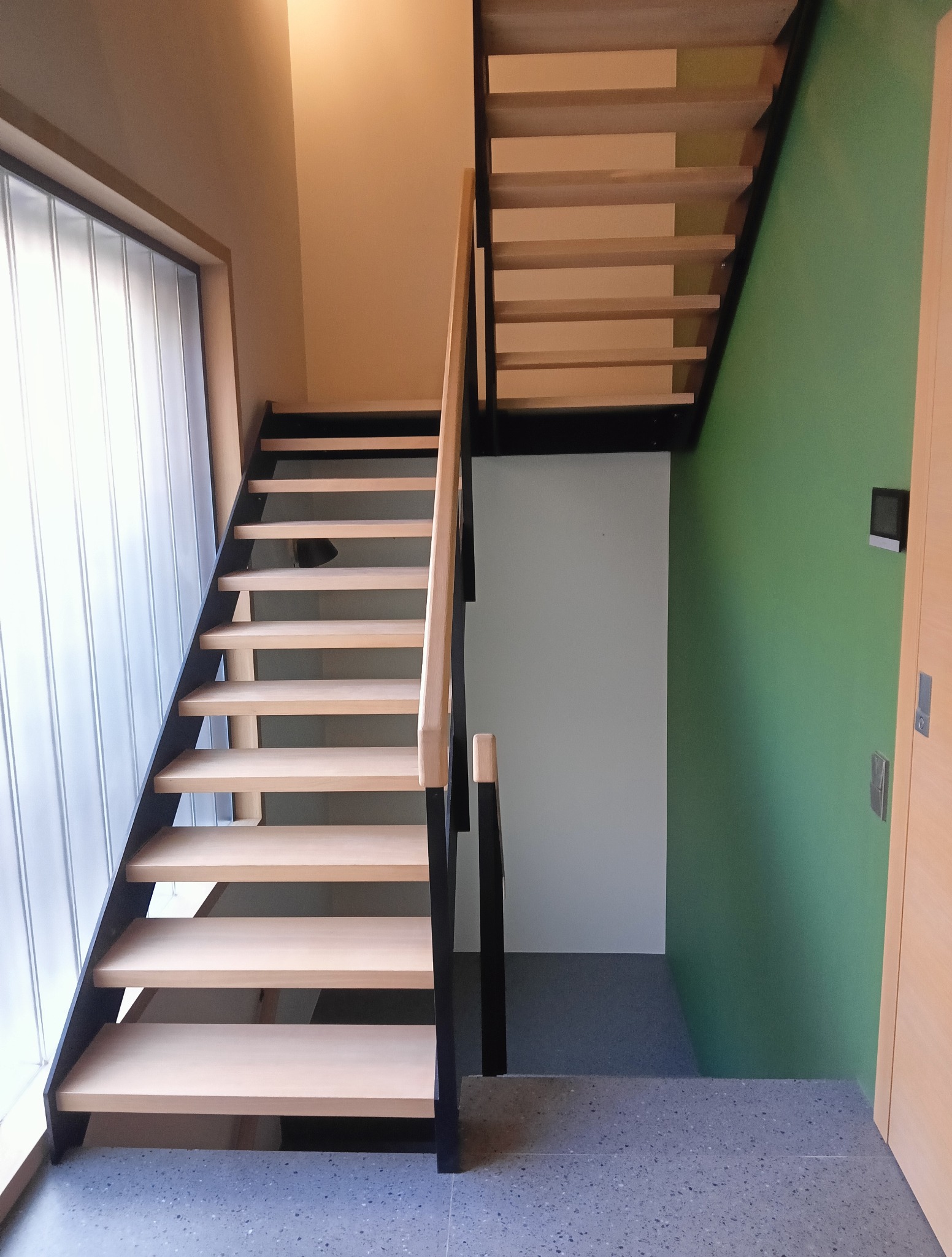 Nowoczesne schody z drewnianymi stopniami i czarną metalową konstrukcją, oświetlone przez okno z luksferami. Minimalistyczny design z zieloną ścianą w tle.