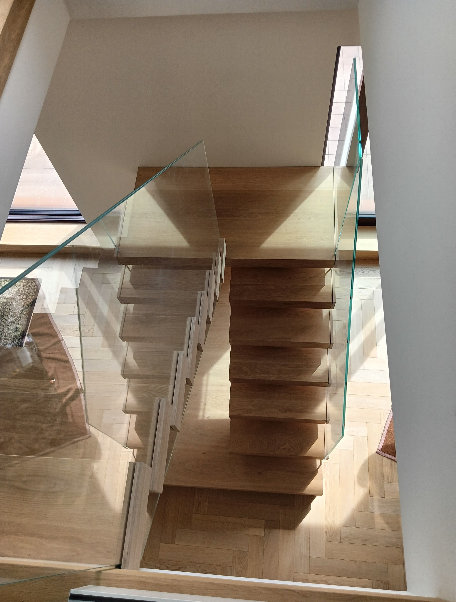 Nowoczesne schody drewniane z szklaną balustradą, widok z góry. Jasne drewno i minimalistyczny design, parkiet ułożony w jodełkę. Doświetlone wnętrze.