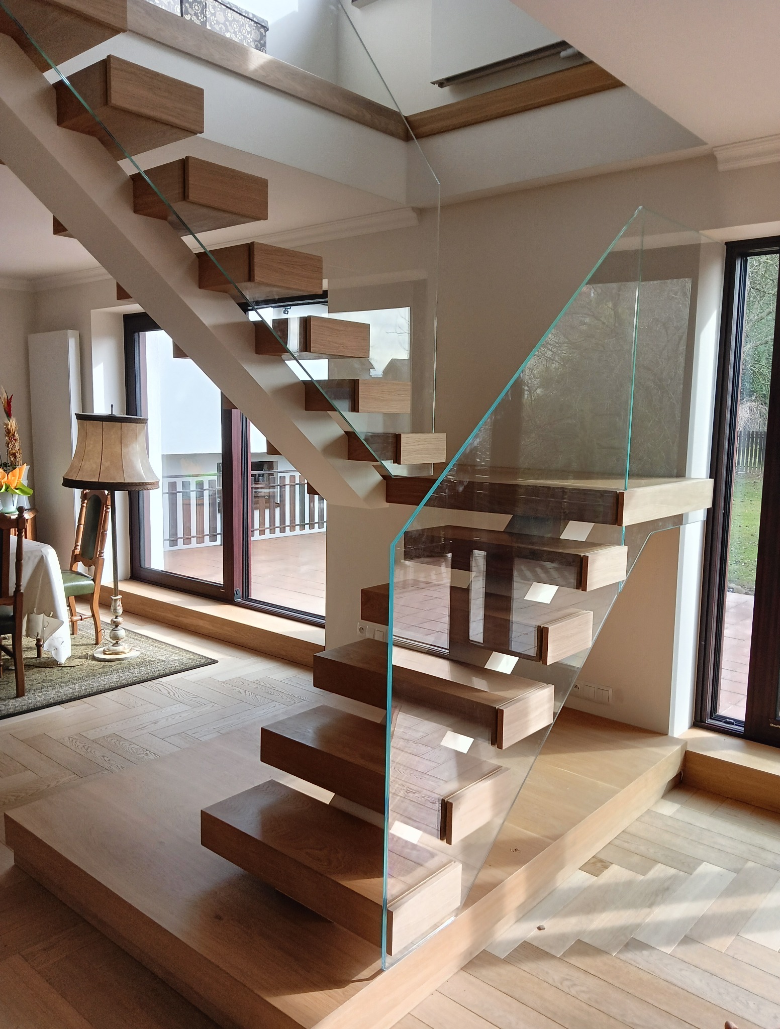 Nowoczesne schody z drewnianymi stopniami wspornikowymi i szklaną balustradą w jasnym wnętrzu z parkietem w jodełkę i widokiem na balkon przez okno.