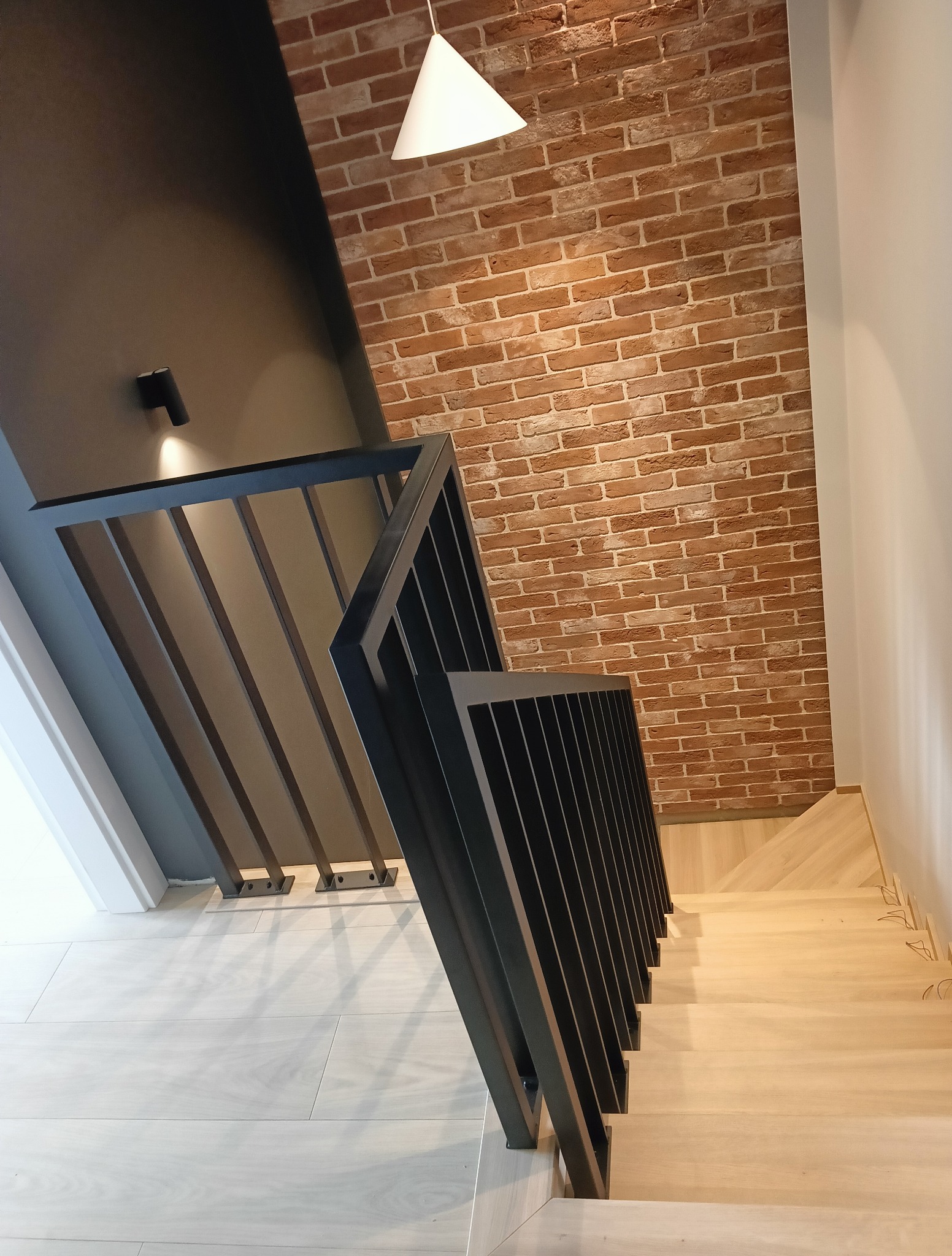 Nowoczesne schody z czarną metalową balustradą i jasnymi drewnianymi stopniami. W tle ściana z czerwonej cegły i designerska lampa wisząca.