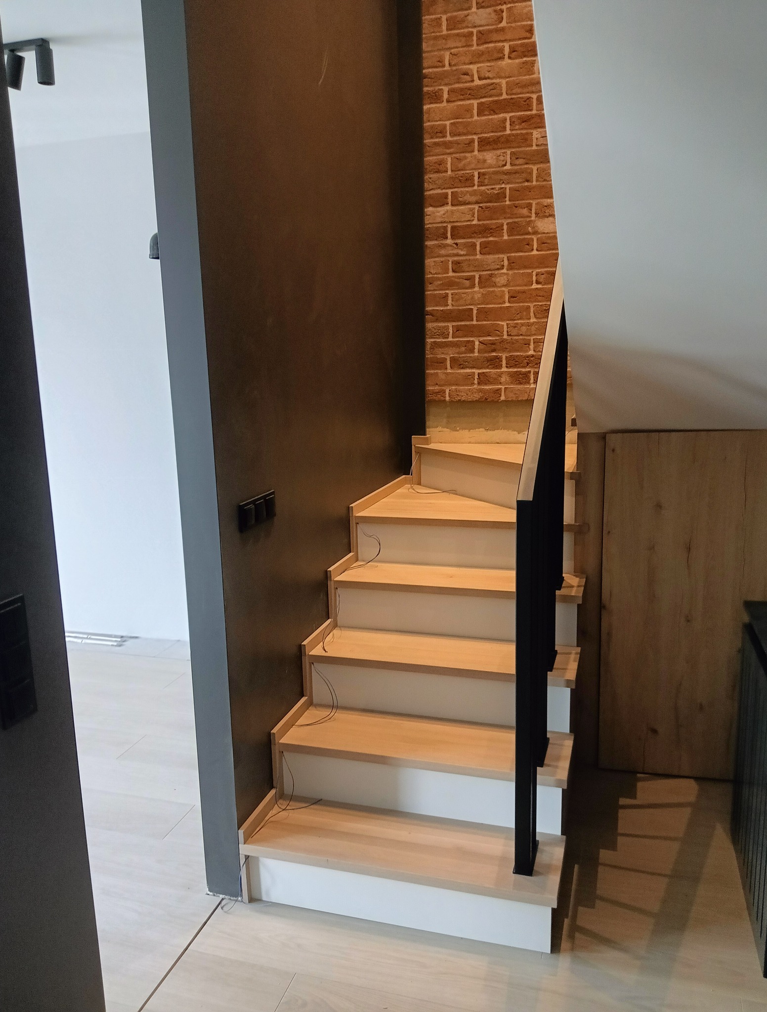 Jasne schody z drewnianymi stopniami i czarną balustradą, osadzone między ścianą z cegły a ciemną ścianą. Widoczne kable przy schodach.