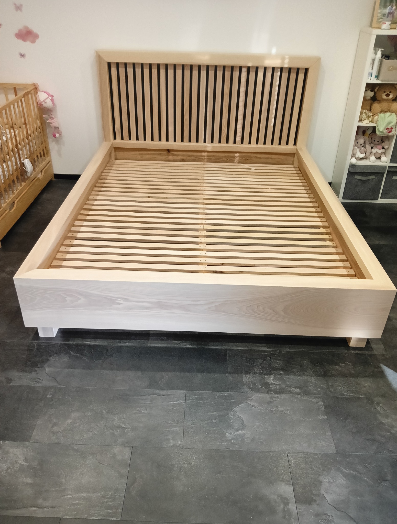 Jasne, drewniane łóżko z listwowym zagłówkiem, ustawione na ciemnej podłodze. Widoczny fragment dziecięcego łóżeczka i regał z zabawkami w tle.