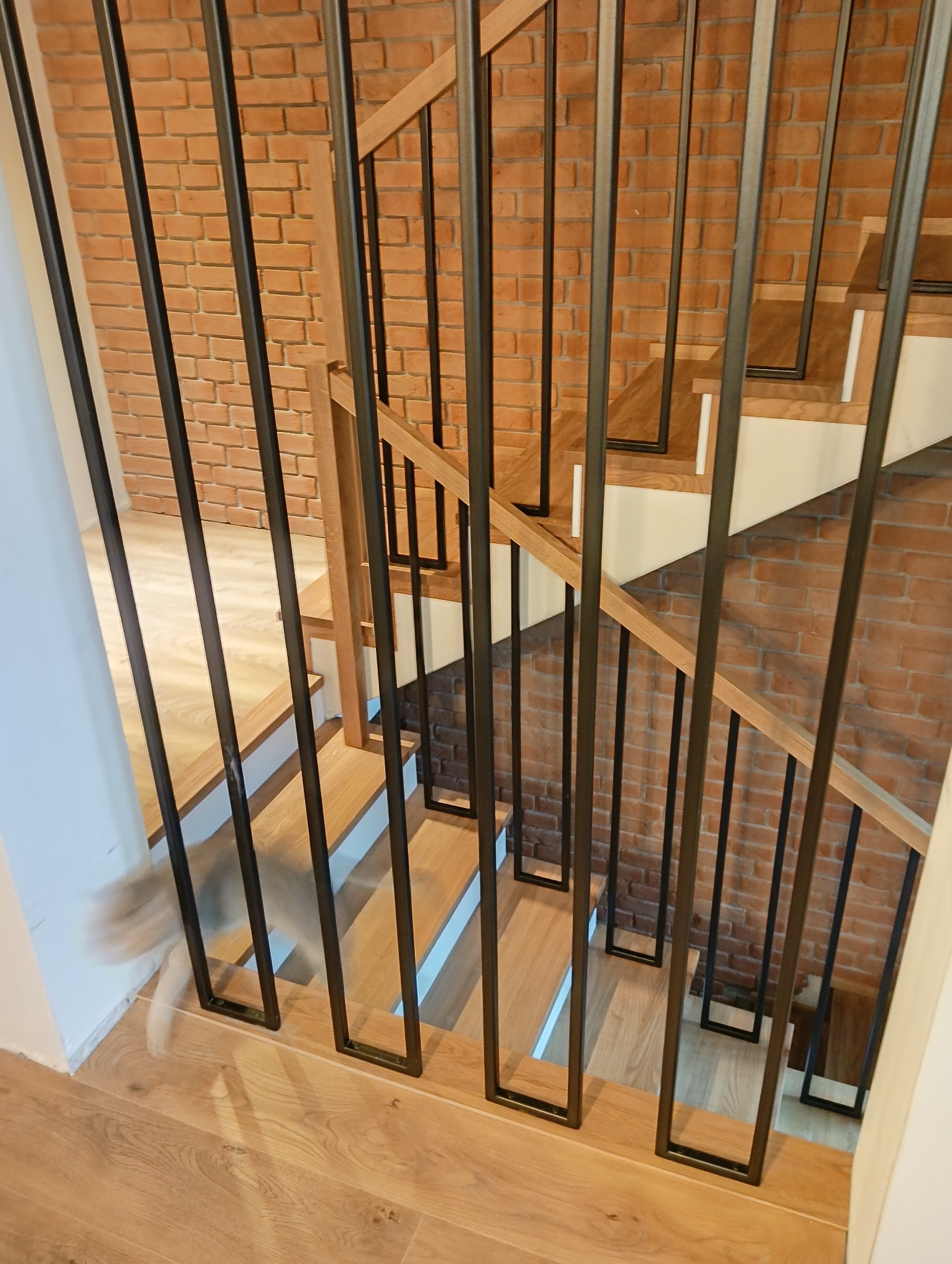 Drewniane schody z czarną, metalową balustradą na tle ściany z cegły. Widoczny fragment podłogi z paneli i przebiegający kot. Ujęcie z góry.