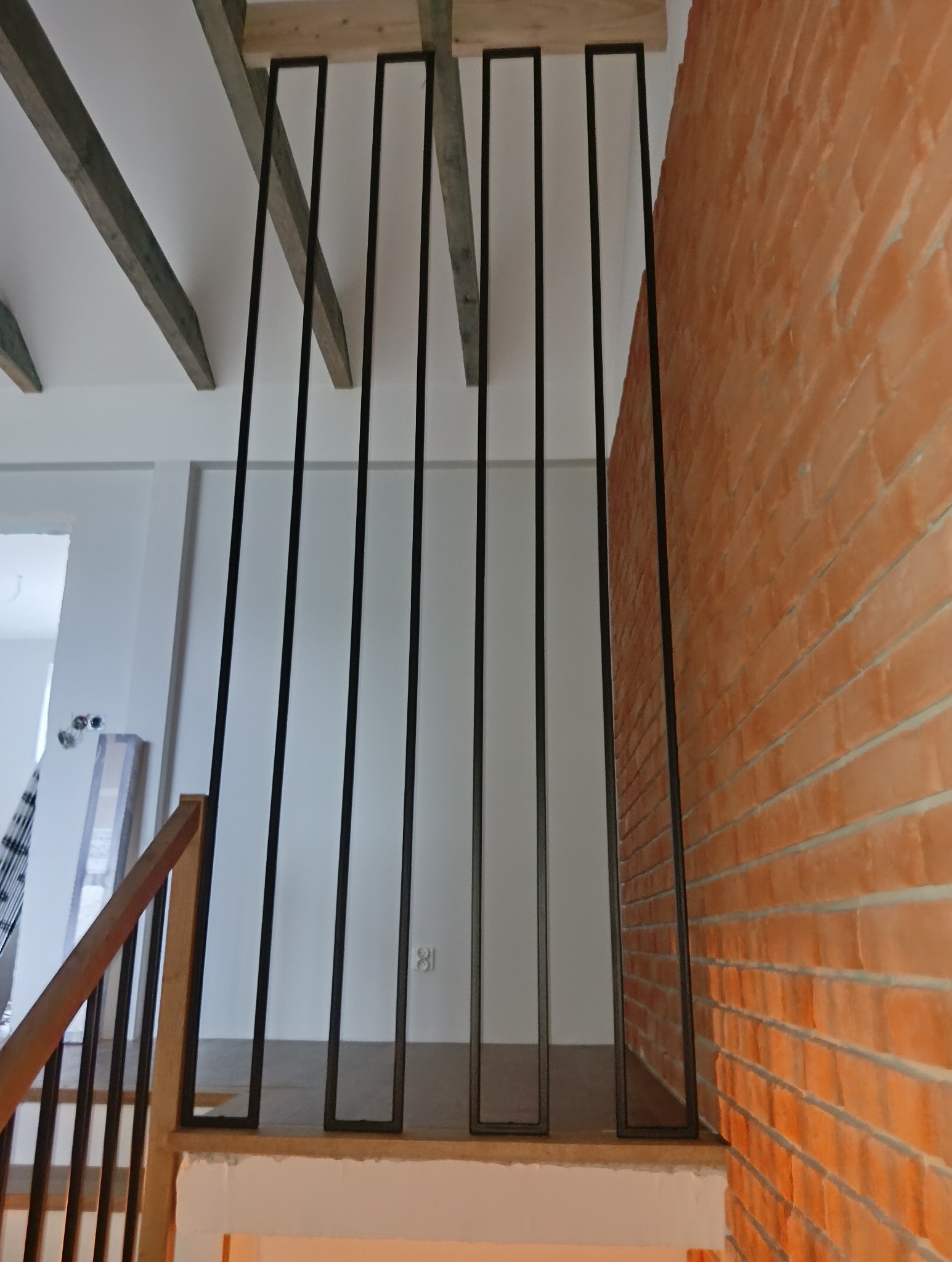 Nowoczesna balustrada metalowa z pionowymi szczeblami na schodach, z ceglaną ścianą w tle i drewnianymi elementami konstrukcyjnymi.