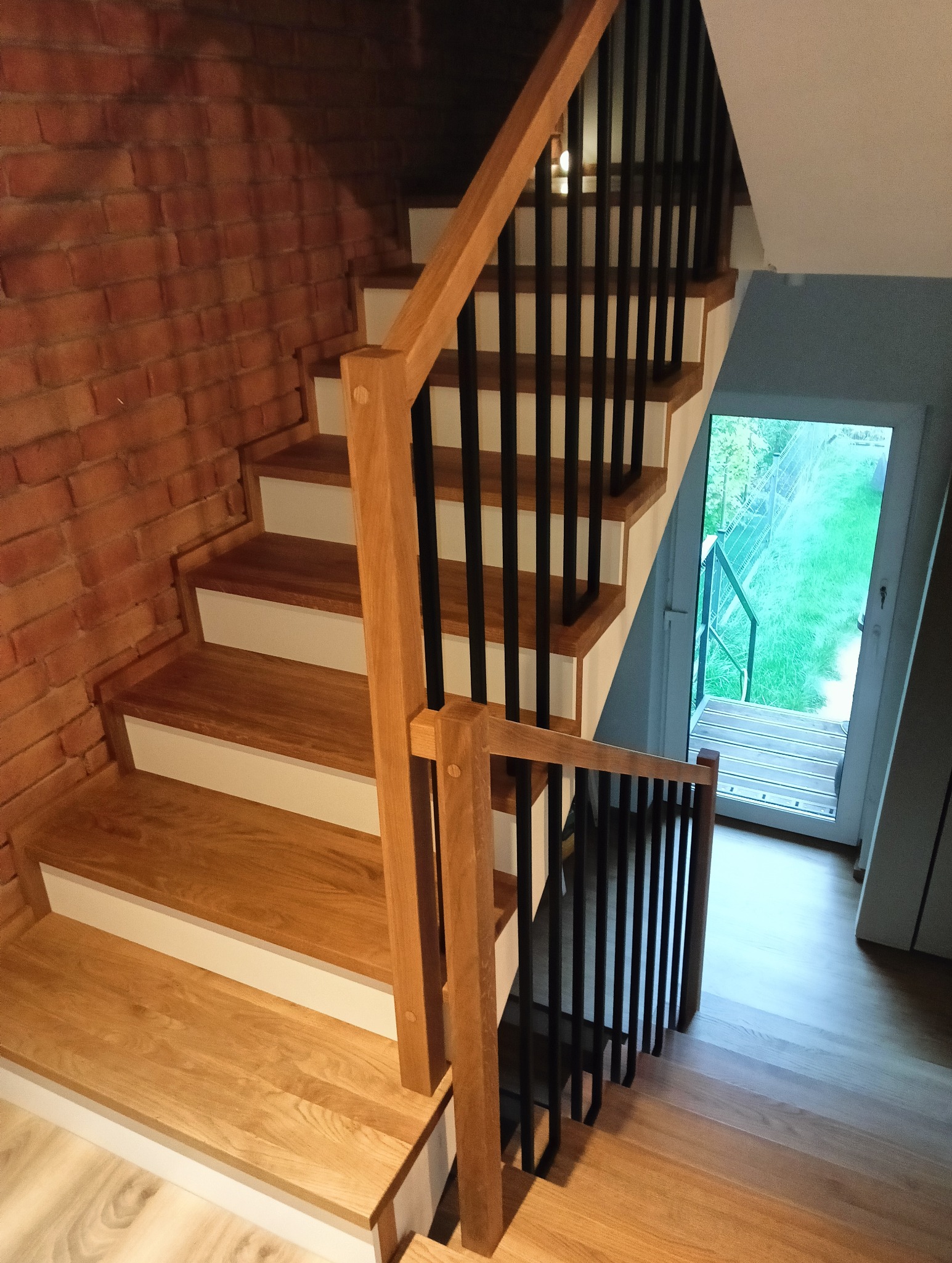 Nowoczesne schody drewniane z czarną balustradą na tle ceglanej ściany. Widok z góry na dół, z widocznym wyjściem na zewnątrz. Jasne drewno i minimalistyczny design.