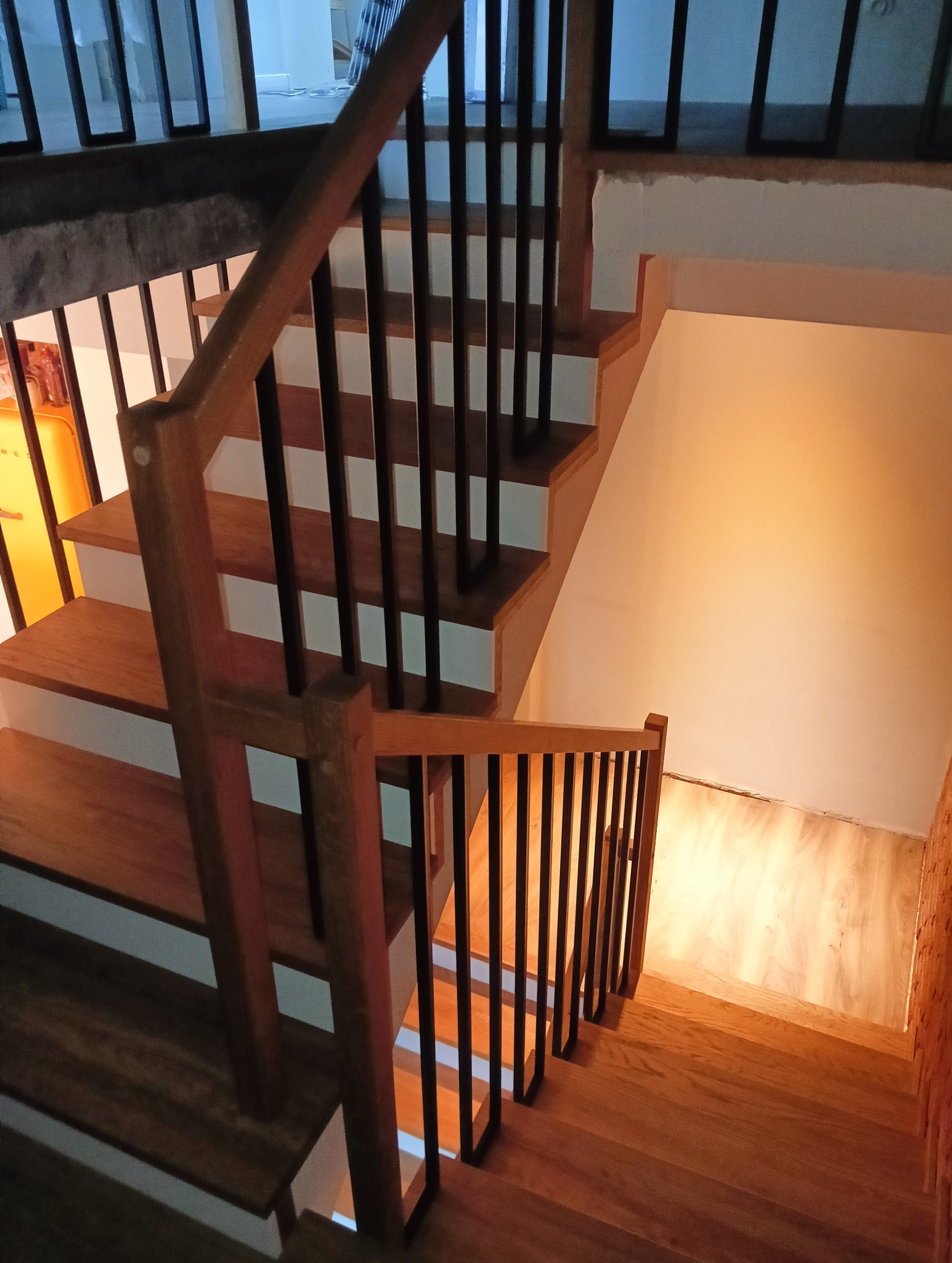Kątowe ujęcie schodów z drewnianymi stopniami i balustradą z czarnymi, pionowymi szczeblami. Widok z góry na dół, oświetlenie podkreśla ciepły odcień drewna.
