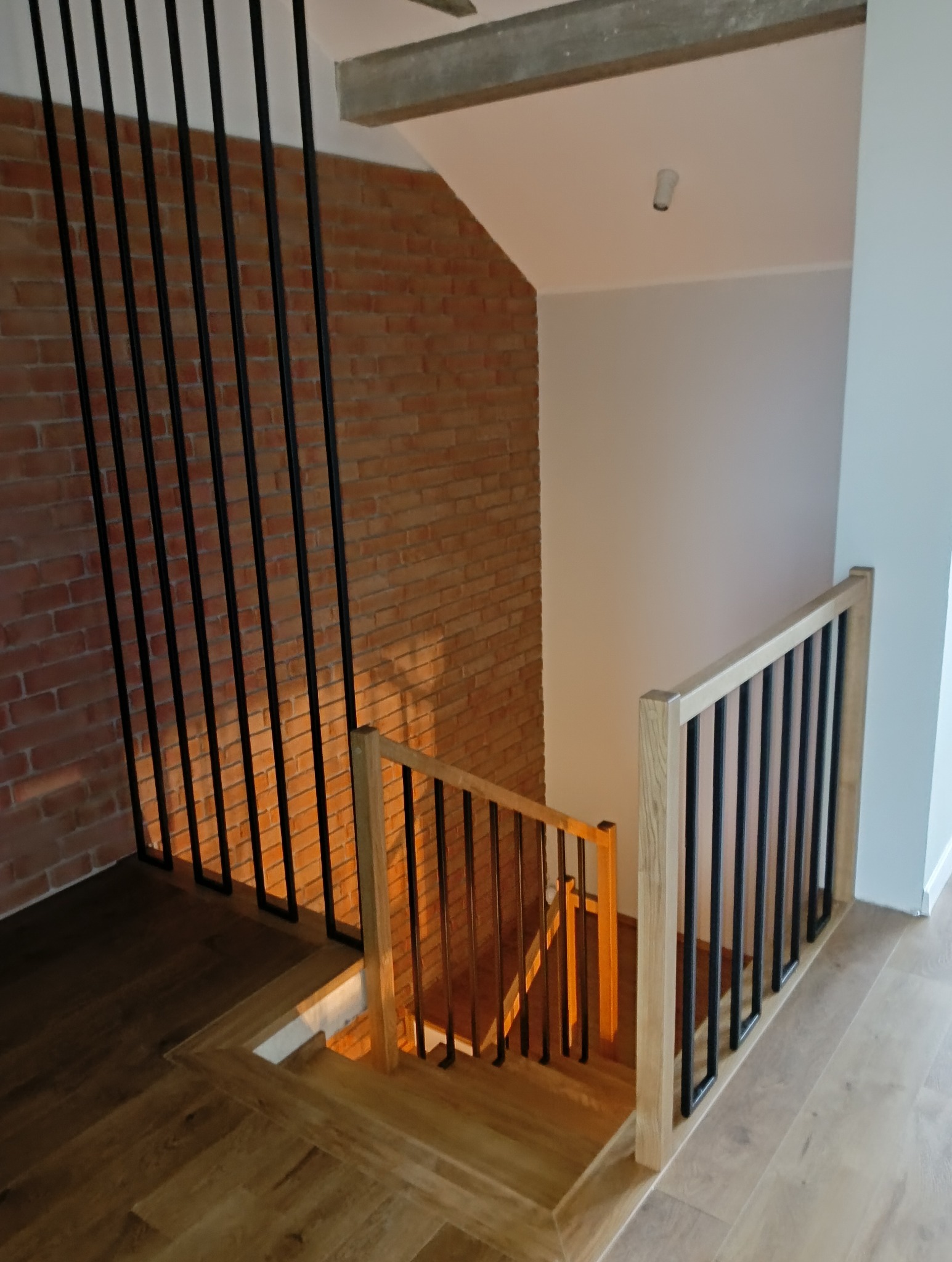 Nowoczesne schody drewniane z czarną, metalową balustradą na tle ceglanej ściany. Jasne drewno schodów kontrastuje z ciemnymi elementami balustrady.