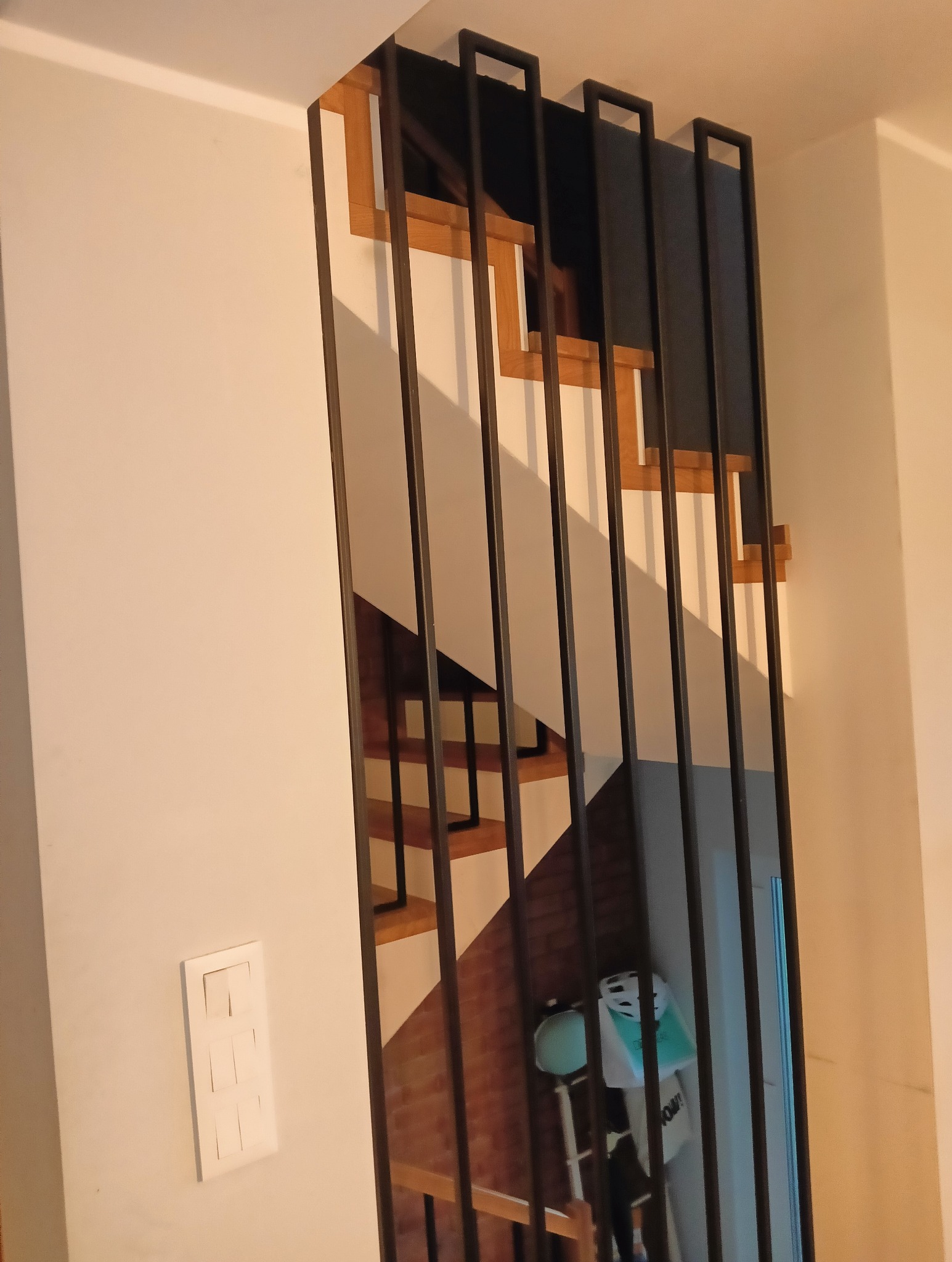 Nowoczesna balustrada metalowa z pionowymi prętami, przez którą widać fragment drewnianych schodów. Minimalistyczny design, jasne wnętrze, widoczny włącznik światła.
