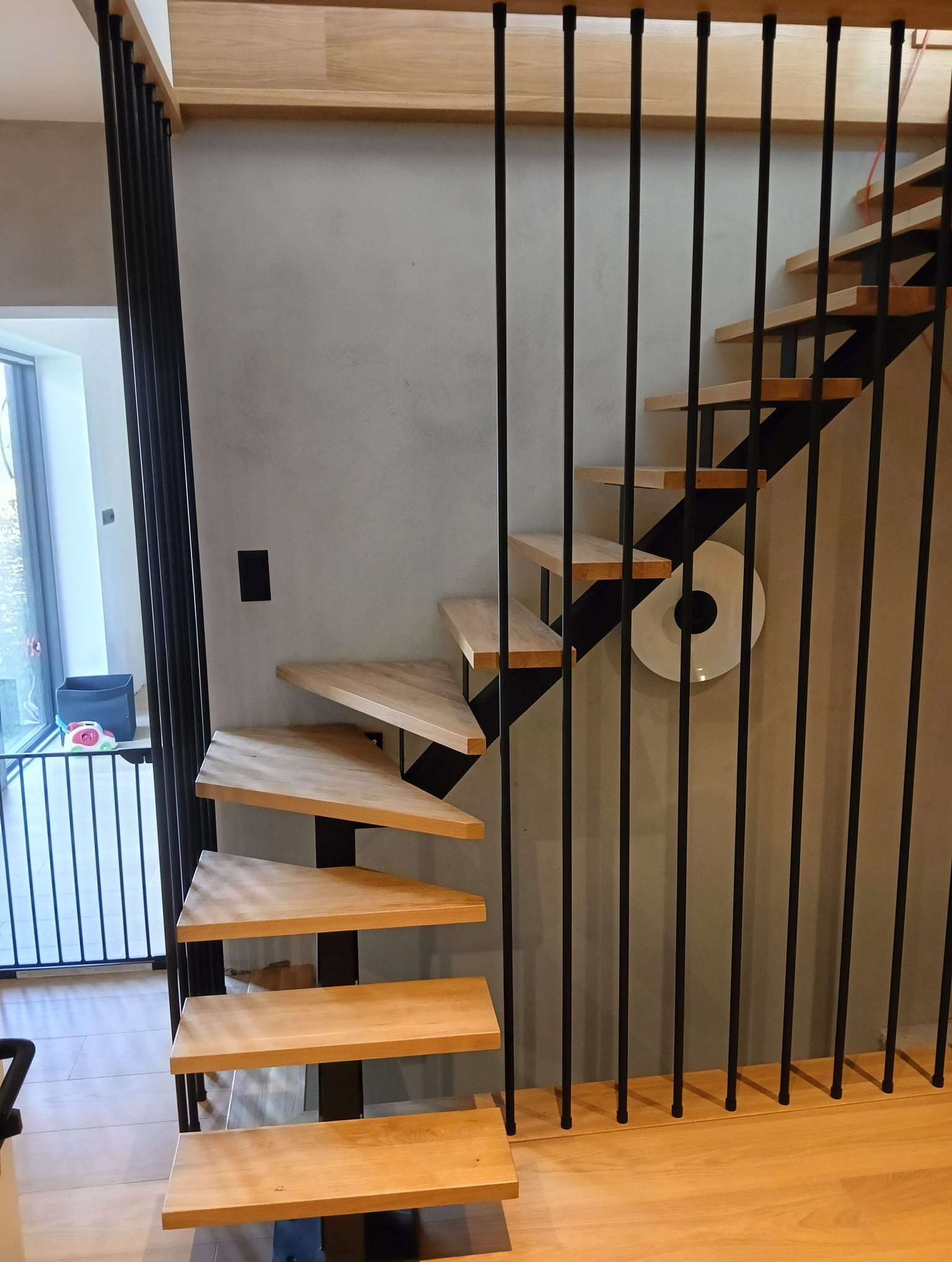 Nowoczesne schody z drewnianymi stopniami i czarną, minimalistyczną balustradą. Widoczne detale wykończenia i oświetlenie boczne.