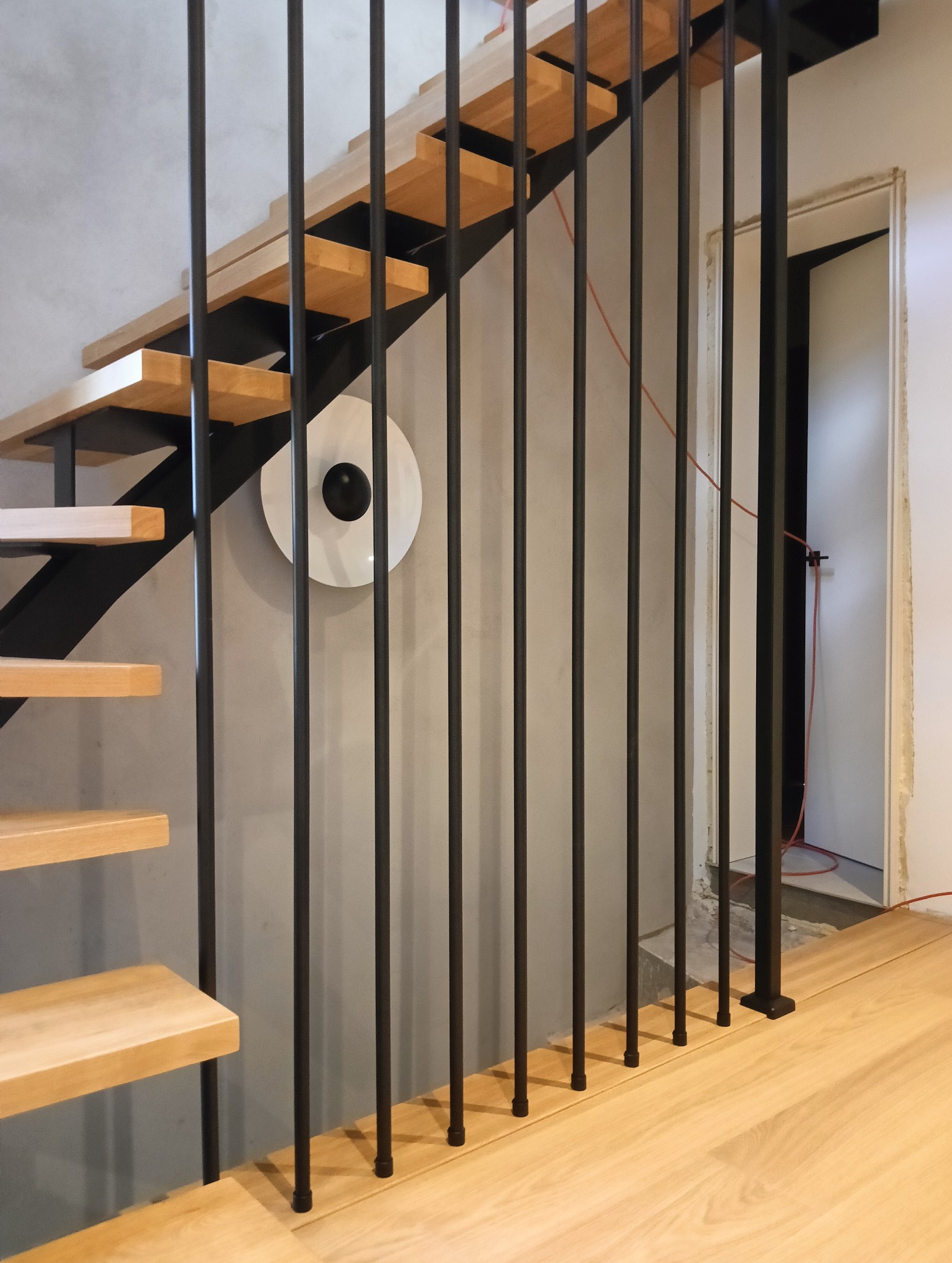Nowoczesne schody z drewnianymi stopniami i czarną, metalową balustradą z pionowych prętów. Na ścianie okrągła lampa. Widok na fragment podłogi i otwarte drzwi.