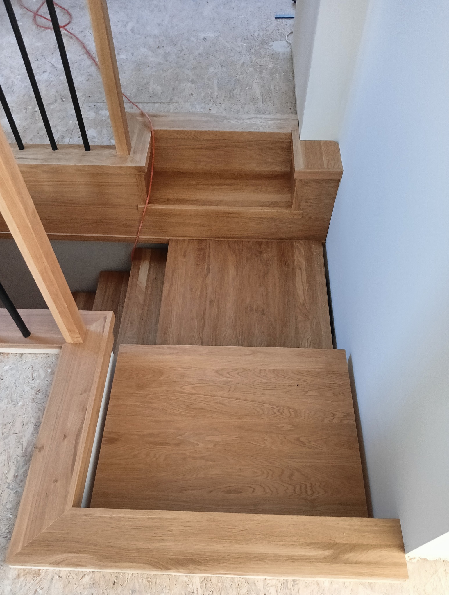 Widok z góry na jasne, dębowe schody z czarnymi, metalowymi balustradami. Nowoczesny design, naturalne drewno, minimalistyczne wykończenie przy białej ścianie.