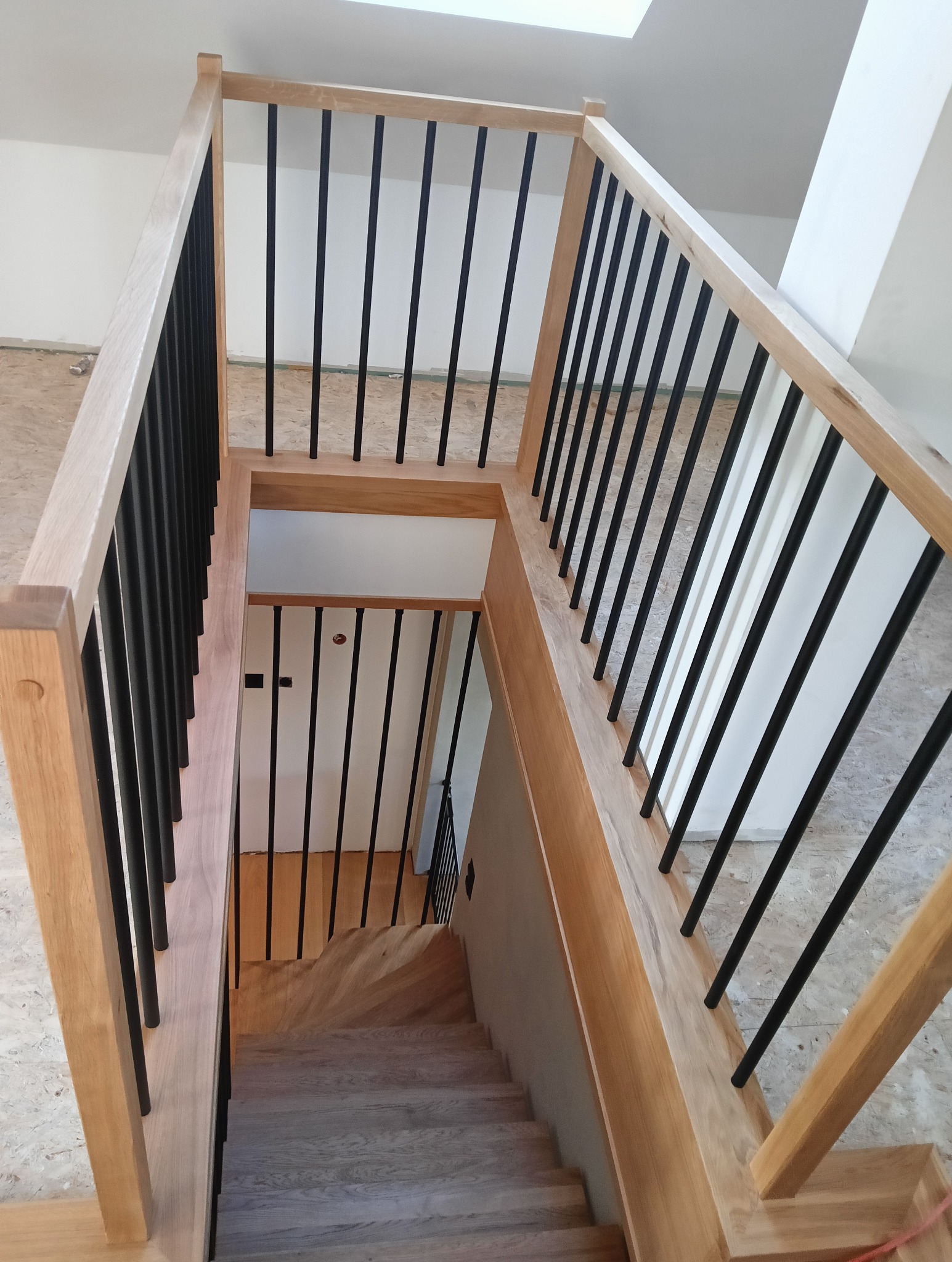 Jasne drewniane schody z czarnymi, pionowymi balustradami, widok z góry na dół, z fragmentem białej ściany i sufitu z oknem dachowym w tle.
