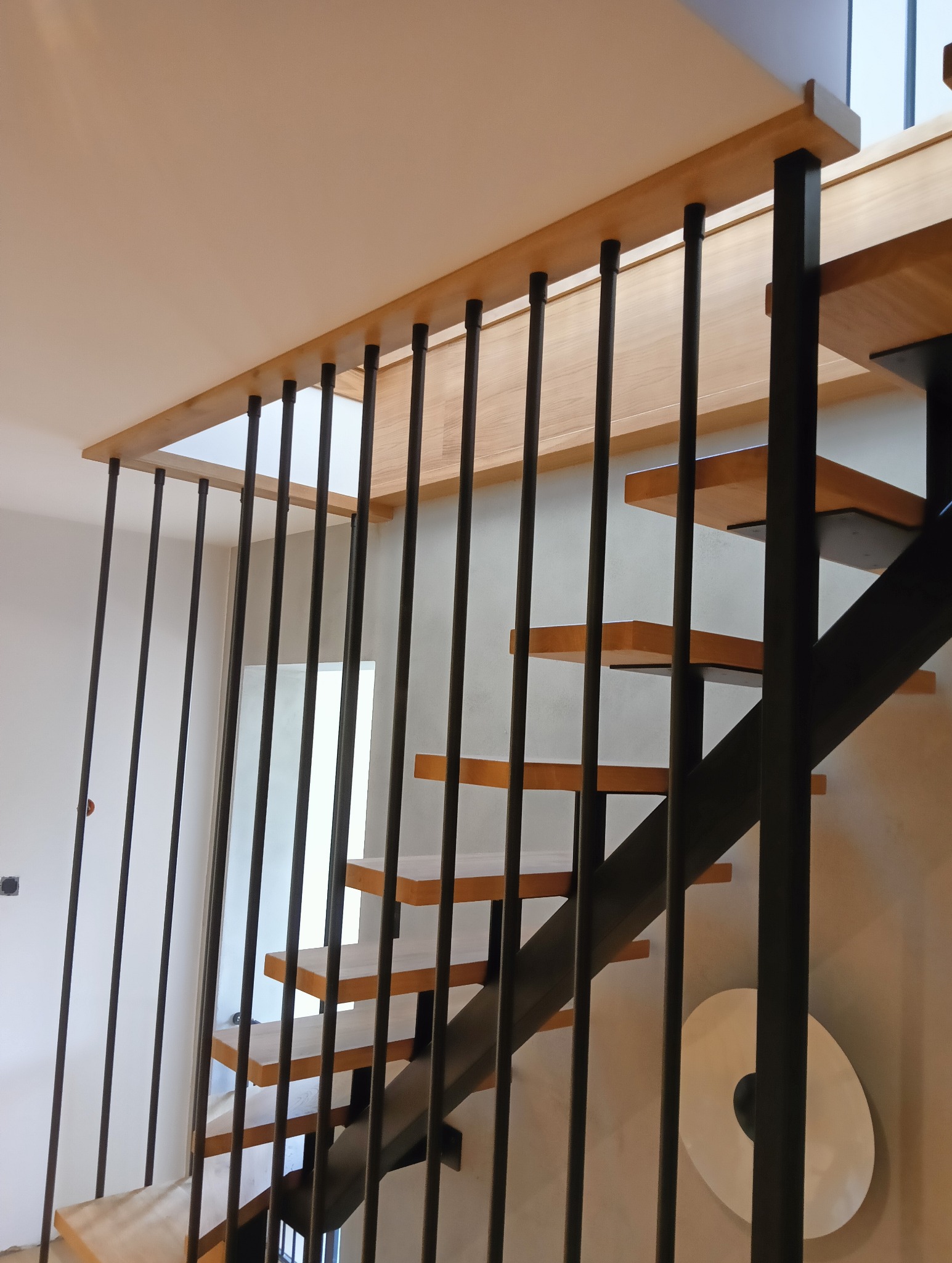 Nowoczesne schody z drewnianymi stopniami i czarną, metalową balustradą. Jasne wnętrze, minimalistyczny design. Widoczne detale mocowania stopni.