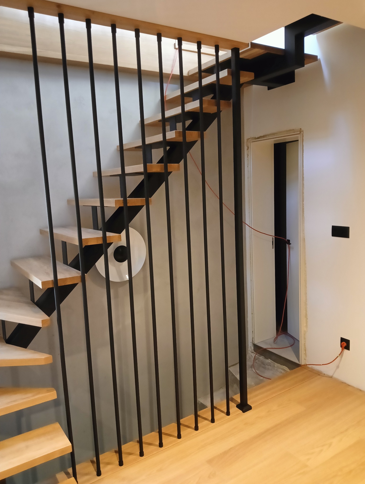 Nowoczesne schody z drewnianymi stopniami i czarną, metalową balustradą w minimalistycznym wnętrzu. Widoczne detale wykończenia i oświetlenie.