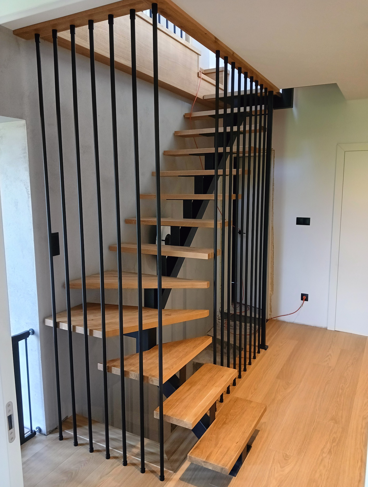 Nowoczesne schody z drewnianymi stopniami i czarną, metalową balustradą z pionowych prętów. Jasne drewno, minimalistyczny design, widok z dołu.