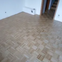 Jasna, nowa podłoga z parkietu ułożonego w kwadraty, widok na białe ściany, narożnik pokoju i schody w tle. Parkiet świeżo po lakierowaniu.