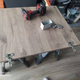 JR- Podłogi Jarosław Rączkowski - Płyta meblowa z zamontowanymi zawiasami, wiertarka akumulatorowa i pudełko z okuciami na wierzchu. Przygotowanie do montażu.