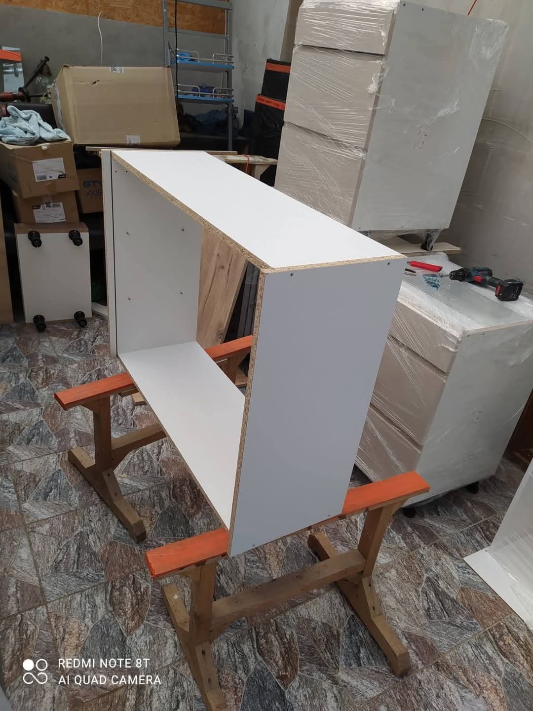 Biały, niedokończony moduł szafki na drewnianym stojaku. W tle kartony i meble w folii. W pobliżu wkrętarka i akcesoria. Widoczne detale montażowe.