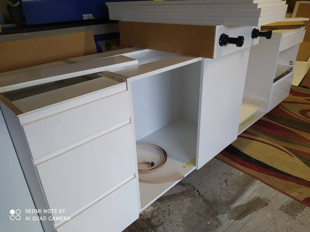 Białe, częściowo zmontowane meble z szufladami i półkami, widoczne uchwyty, elementy montażowe i podłoga. Meble w trakcie montażu.