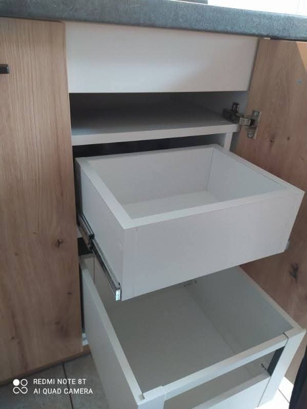 Szafka kuchenna z dwoma wysuwanymi, białymi szufladami na metalowych prowadnicach. Fronty szafki z jasnego drewna. Widoczne zawiasy i blat.