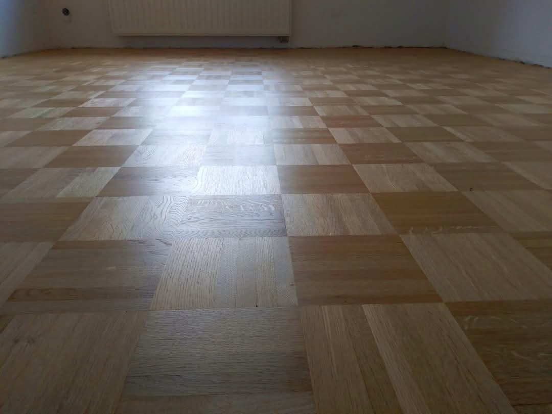 Parkiet w jasnym odcieniu drewna ułożony w kwadraty, widok z perspektywy podłogi, z widocznym grzejnikiem na ścianie. Szlifowana powierzchnia odbija światło.