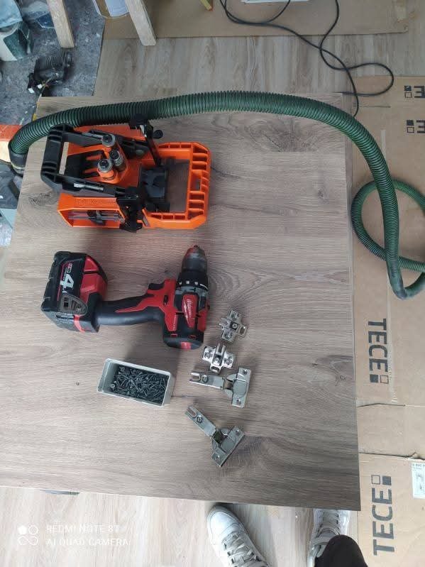 Wiertarka, zawiasy meblowe i wkręty na blacie imitującym drewno, obok karton z napisem TECE i odkurzacz. Przygotowanie do montażu mebli.