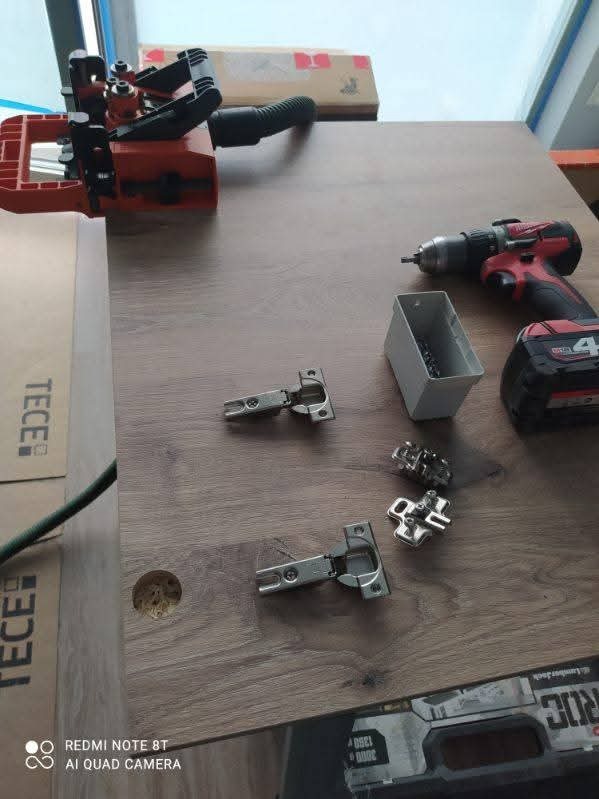 Przygotowanie do montażu zawiasów na drewnianej płycie w Proszowicach. Widoczne narzędzia: wiertarka, wiertła i zawiasy. Otwór pod zawias już wywiercony.