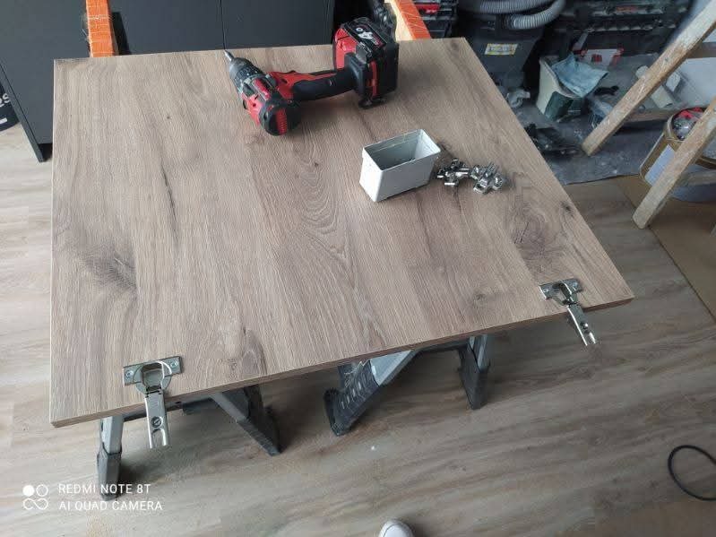 Płyta meblowa z zamontowanymi zawiasami, wiertarka akumulatorowa i pudełko z okuciami na wierzchu. Przygotowanie do montażu.